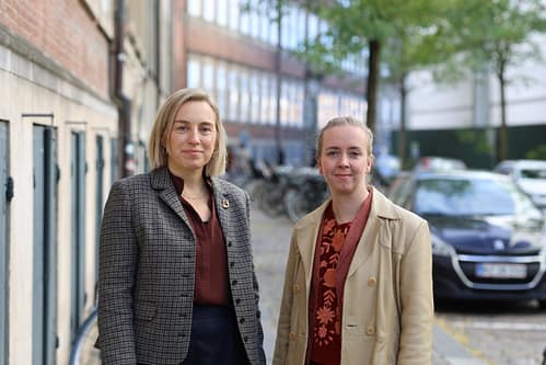Marie Bender Foltmann og Mikkeline Thomsen står side om side på en brostensbelagt gade i København. Marie Bender Foltmann til venstre har kort, lyst hår og er iført en ternet blazer over en mørkerød skjorte. Mikkeline Thomsen til højre har lyst hår sat op og bærer en beige jakke over en rødblomstret bluse. Begge ser smilende mod kameraet. I baggrunden ses byens bygninger, cykler og parkerede biler.